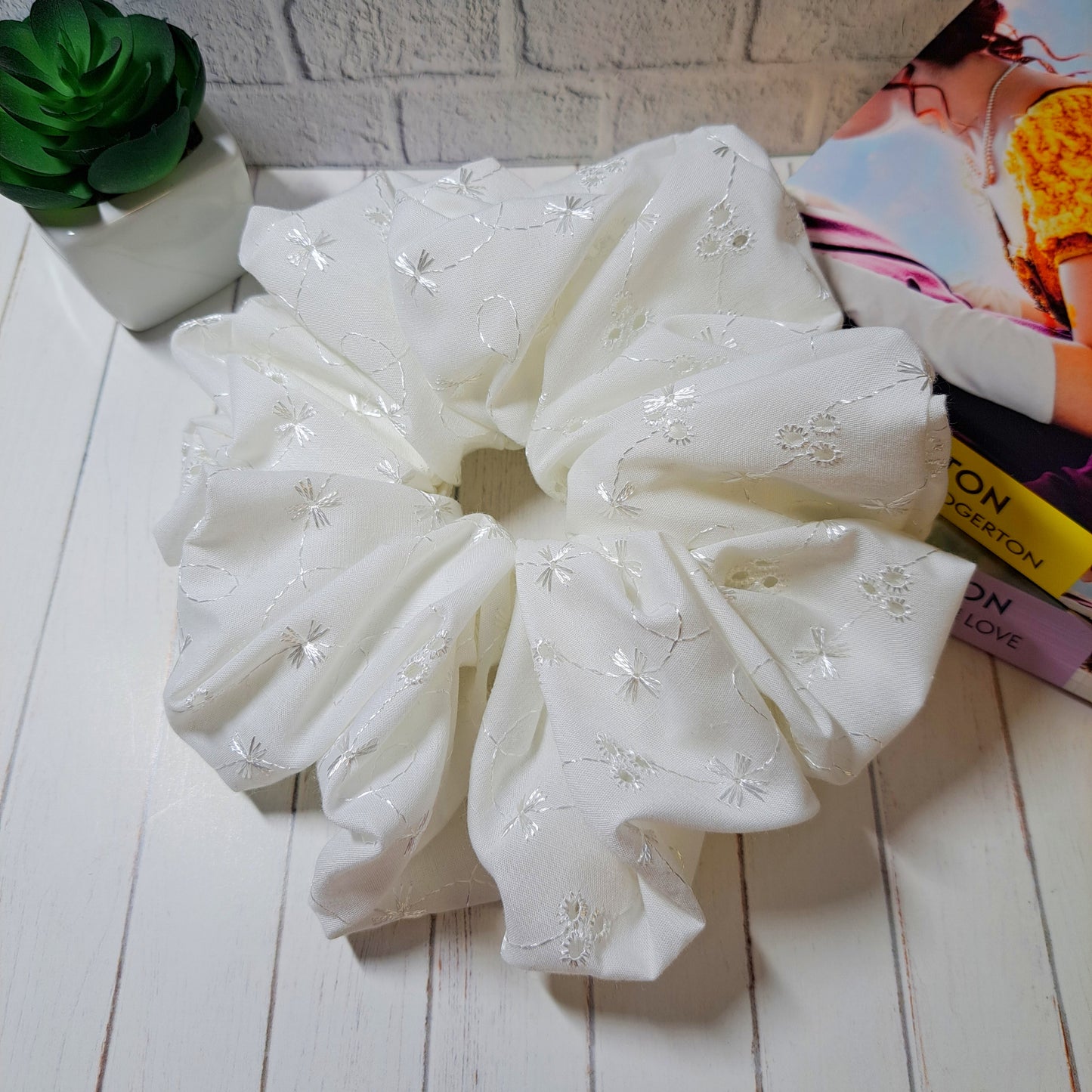 Adaline, our ivory coloured xl sized broderie anglaise scrunchie, displayed on a white wooden slatted background with a small green plant and two Bridgerton Books as decorations