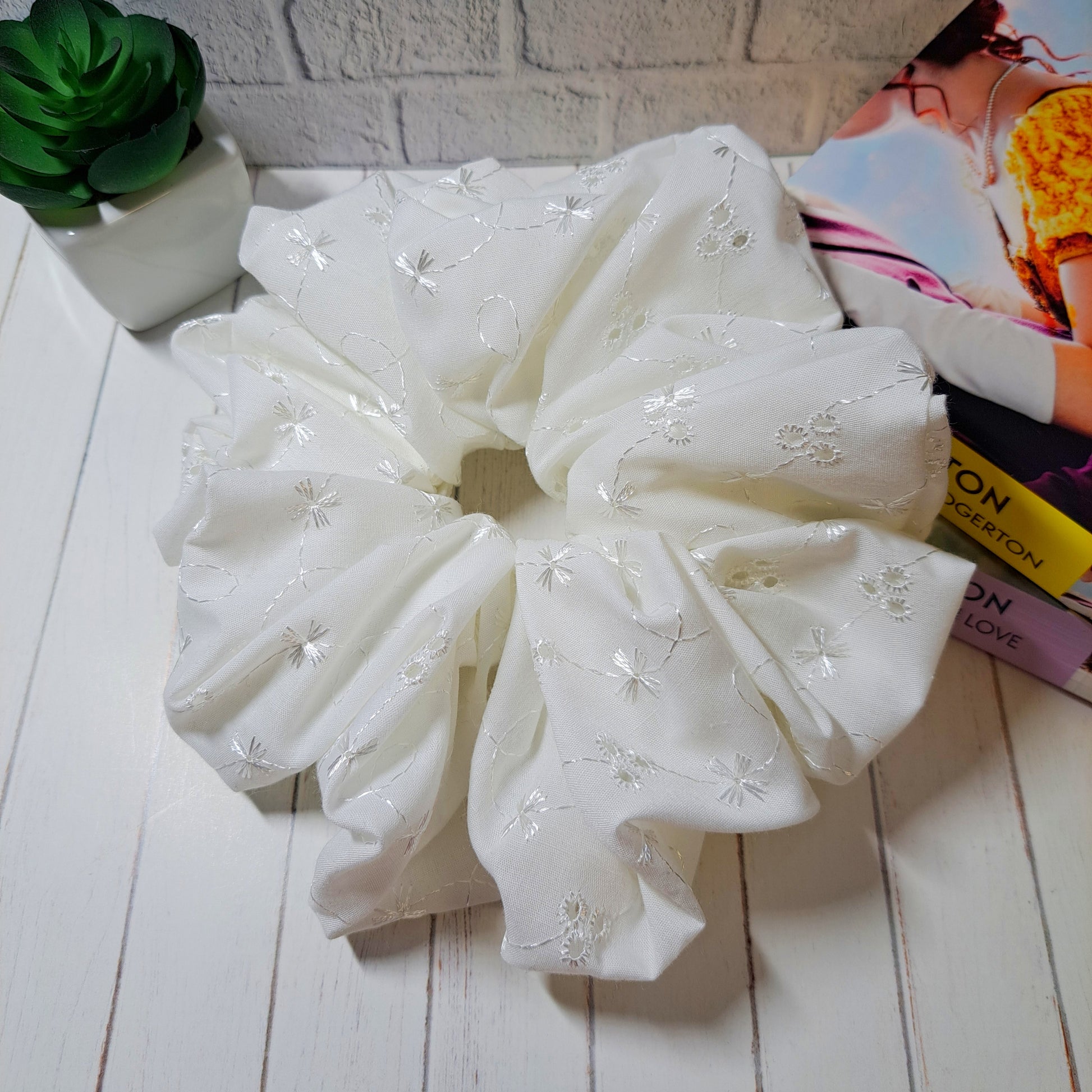 Adaline, our ivory coloured xl sized broderie anglaise scrunchie, displayed on a white wooden slatted background with a small green plant and two Bridgerton Books as decorations