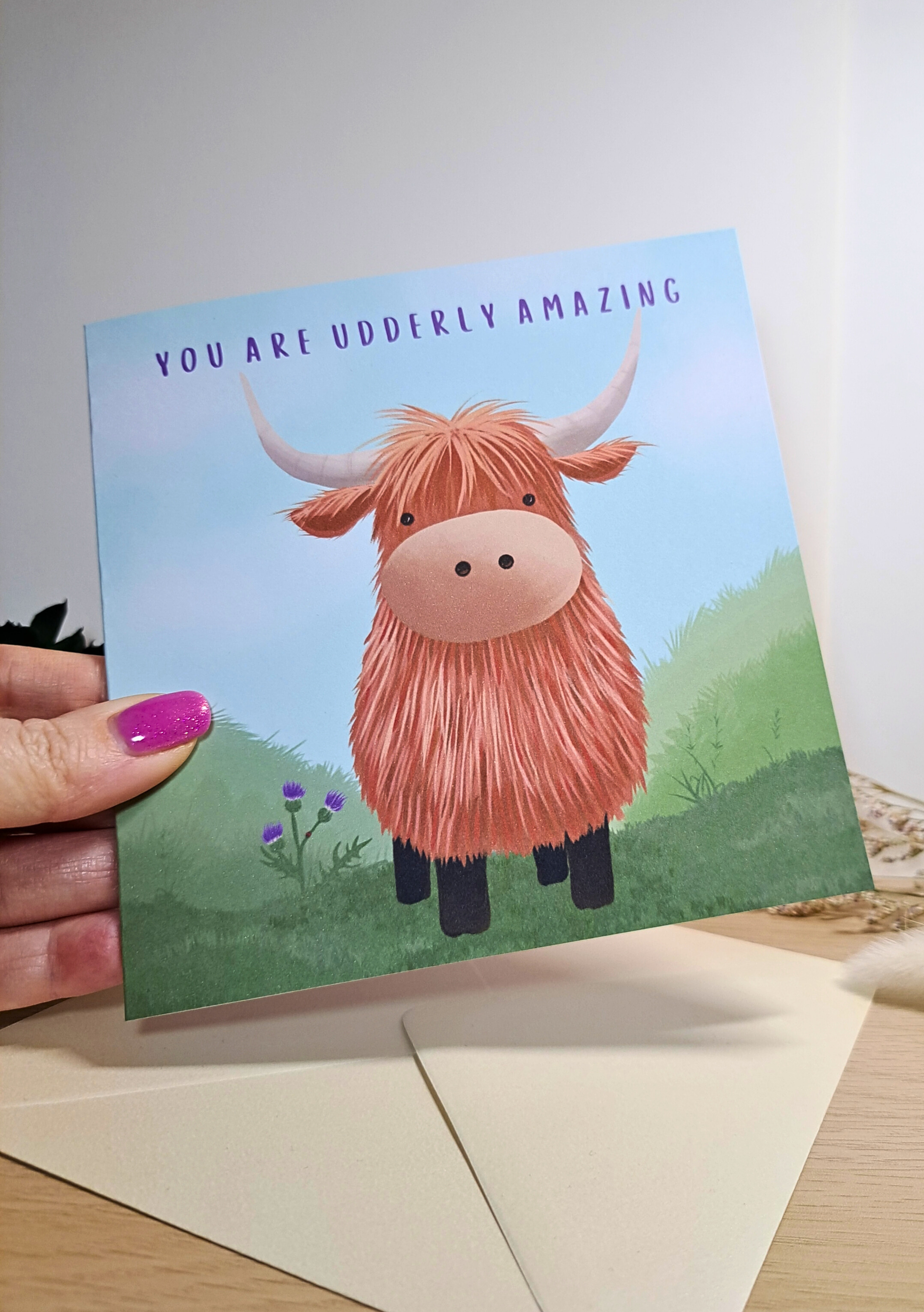 Greeting card with a highland cow illustration and text 'You are udderly amazing' held by a hand.