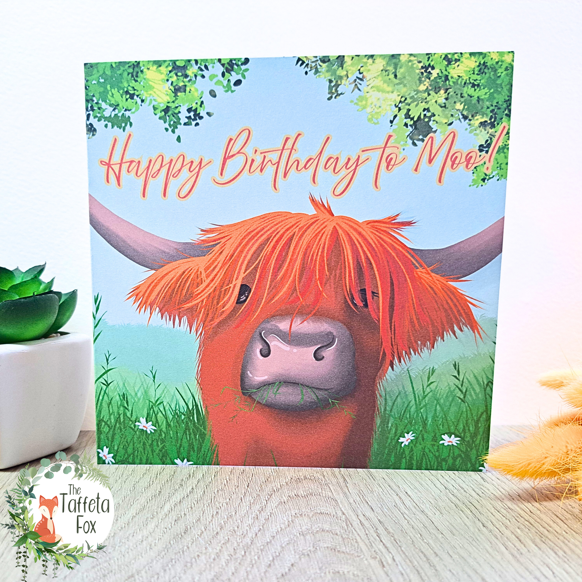 Square birthday card showing an illustrated Highland cow with long ginger fringe and the words “Happy Birthday to Moo!” surrounded by grass and woodland leaves.