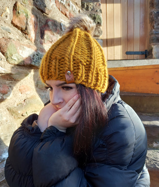 A young lady with long dark hair wearing a mustard coloured chunky hand knitted hat. The hat features a brown faux leather tag and brown pompom.