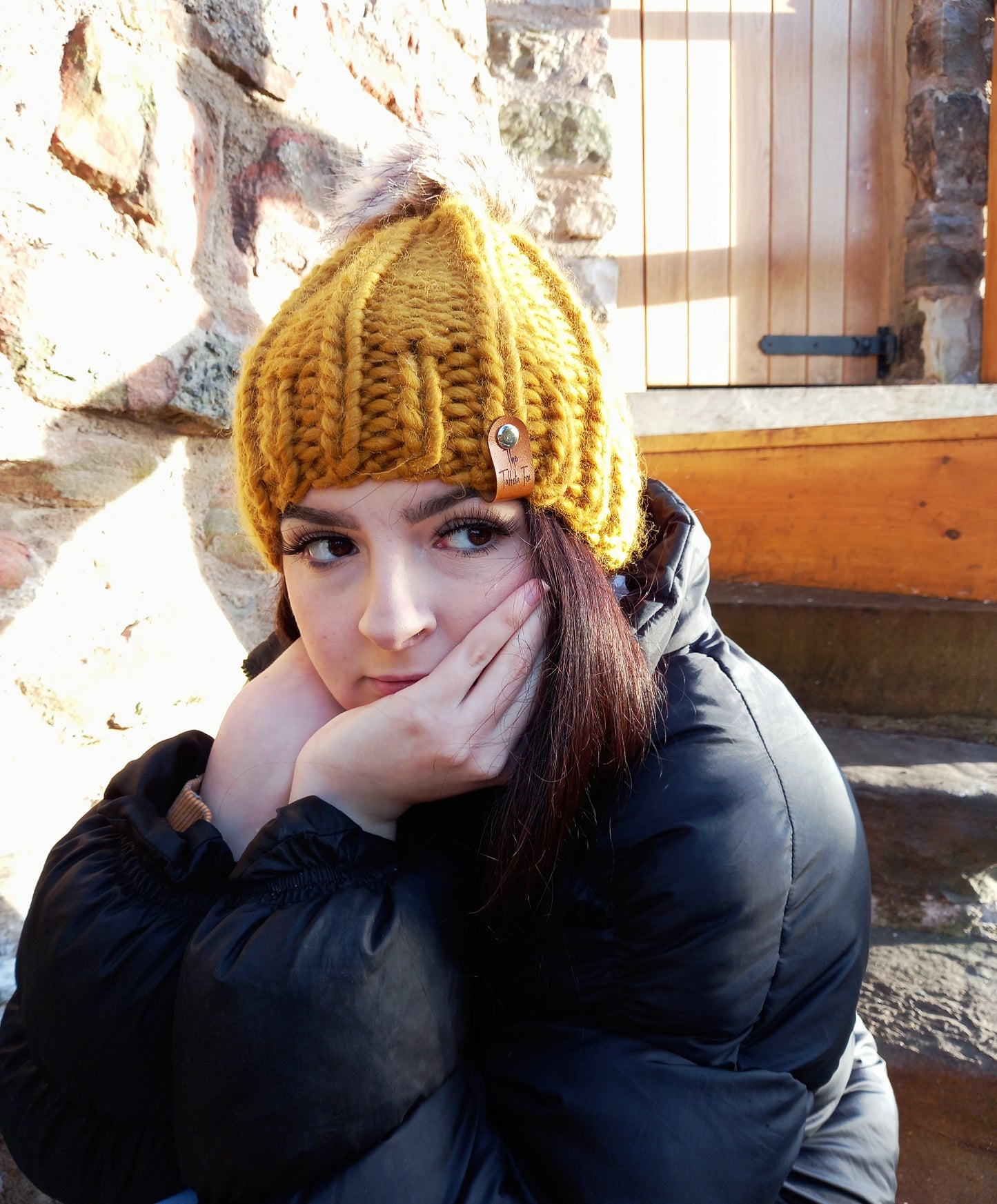 A young lady with long dark hair wearing a mustard coloured chunky hand knitted hat. The hat features a brown faux leather tag and brown pompom.
