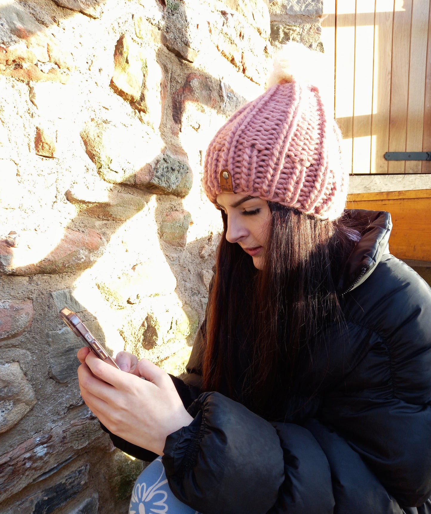 This image shows a young woman with long dark hair wearing a chunky hand knitted hat in pink with a cream pompom.