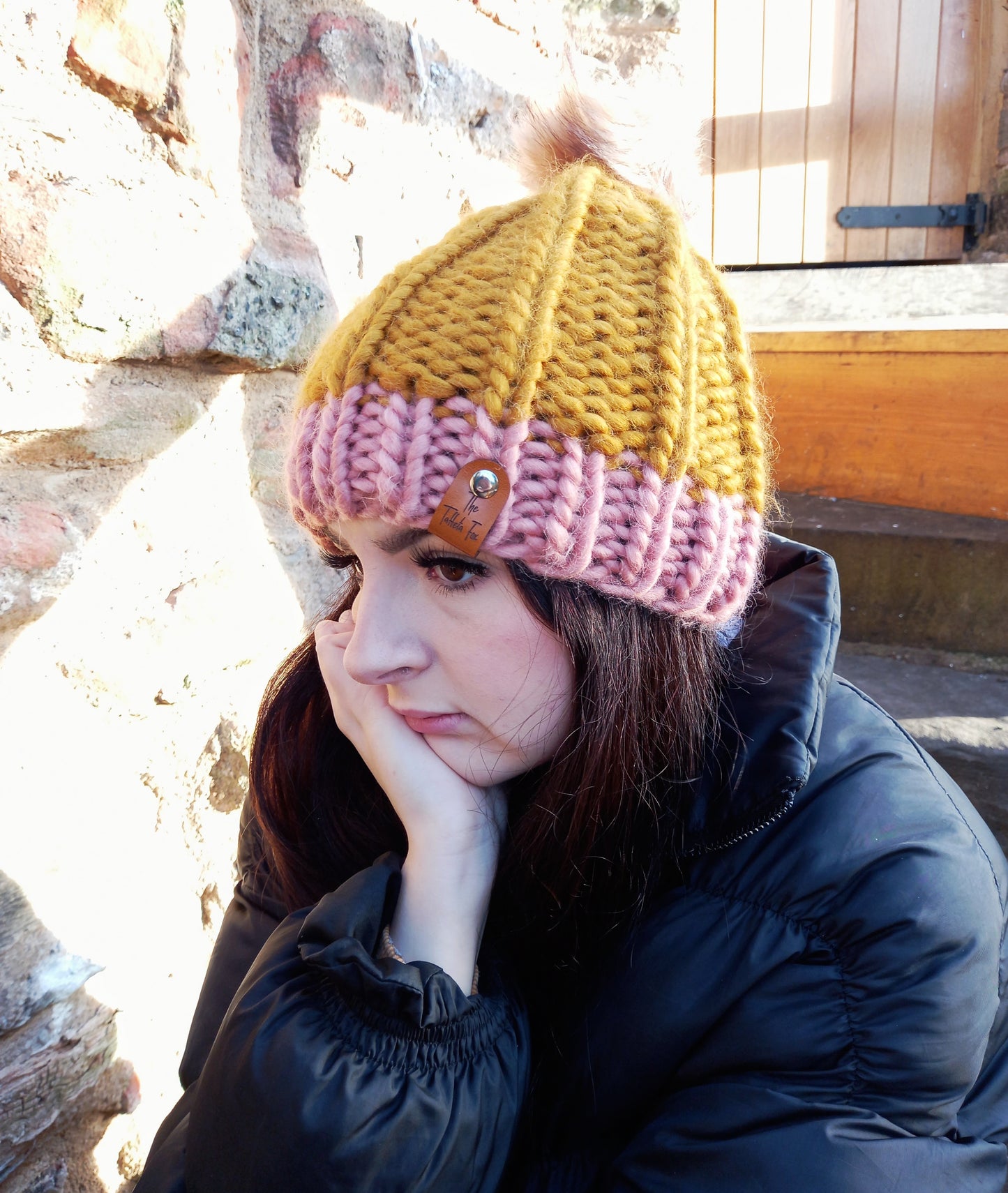 This image shows a super chunky knitted hat in mustard with a pink brim, worn by a young lady with long dark hair. It features a fluffy creamy coloured pompom. It also shows the small brown faux leather Taffeta Fox label attached via a small silver rivet.