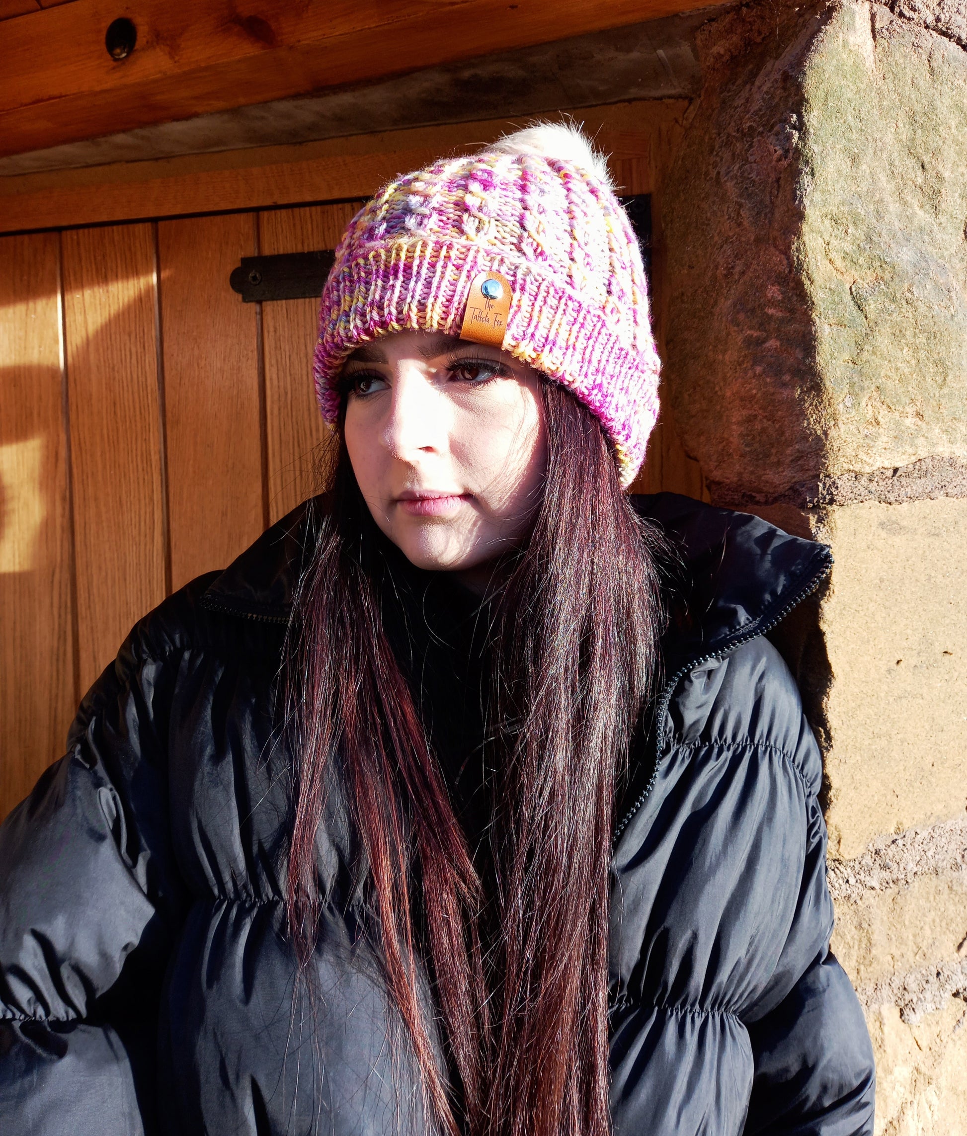 This image shows a young lady with long dark hair wearing a hand knitted hat with a faux cable pattern with a wide fold up brim and is a lovely mixture of pinks, yellows, purples and blues. It features a small faux leather 'Taffeta Fox' label and a soft cream removeable pompom.