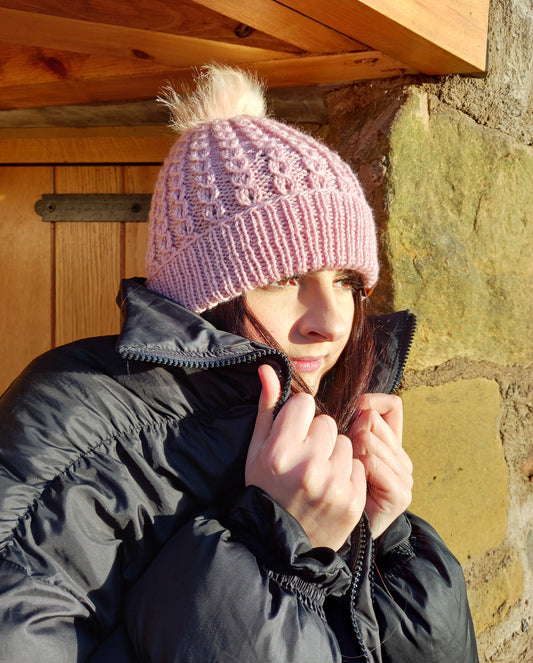 A young lady with long dark hair and black coat wearing a pink hand knitted faux cable patterned hat featuring a creamy coloured pompom.