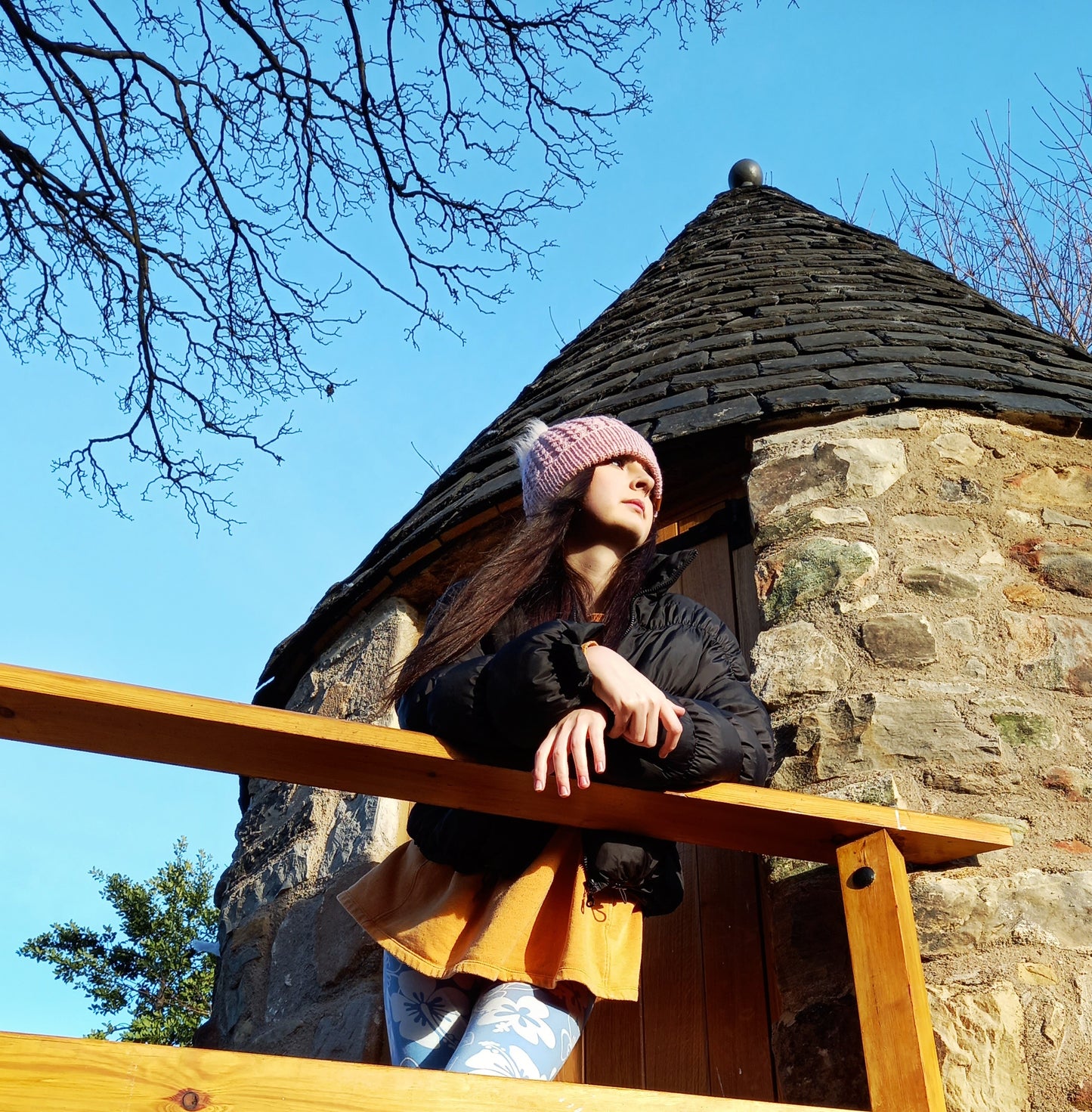 A young lady leaning over the balcony of an old stone tower, wearing a hand knitted pink hat featuring a creamy coloured pompom.