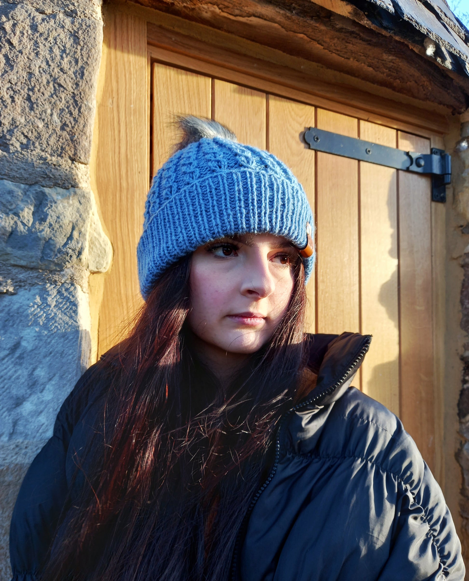 This image shows a young lady with long dark hair wearing a faux cable hand knitted hat with turned up brim in a lovely shade of blue. The hat features a light grey pompom and brown faux leather The Taffeta Fox tag.