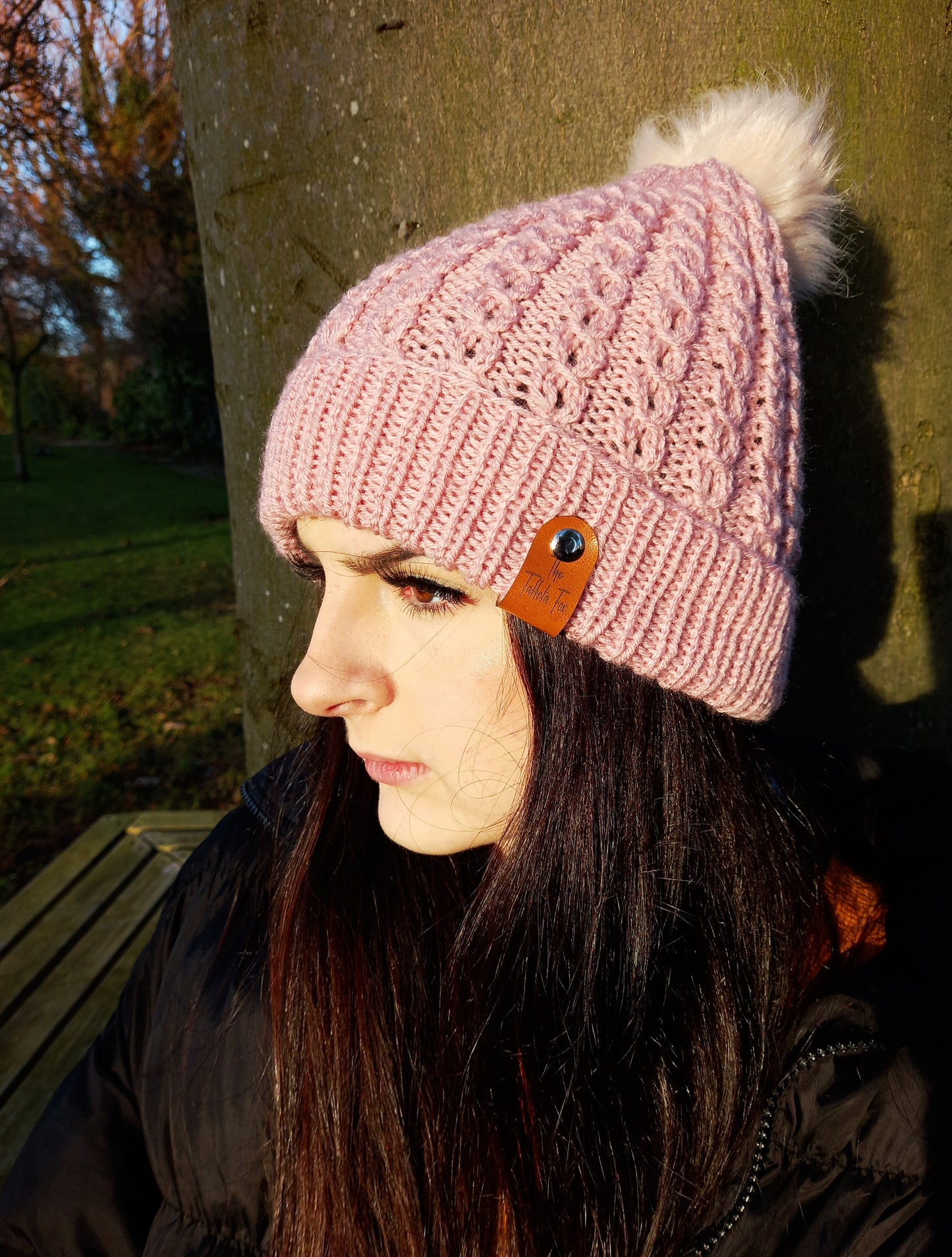 A young lady with long dark hair wearing a hand knitted faux cable patterned hat in a dusky shade of pink. The hat features a brown faux leather tag and creamy coloured pompom.