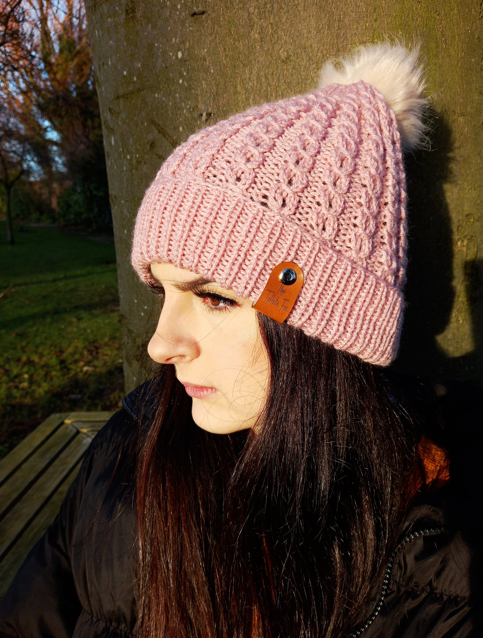 A young lady with long dark hair wearing a hand knitted faux cable patterned hat in a dusky shade of pink. The hat features a brown faux leather tag and creamy coloured pompom.