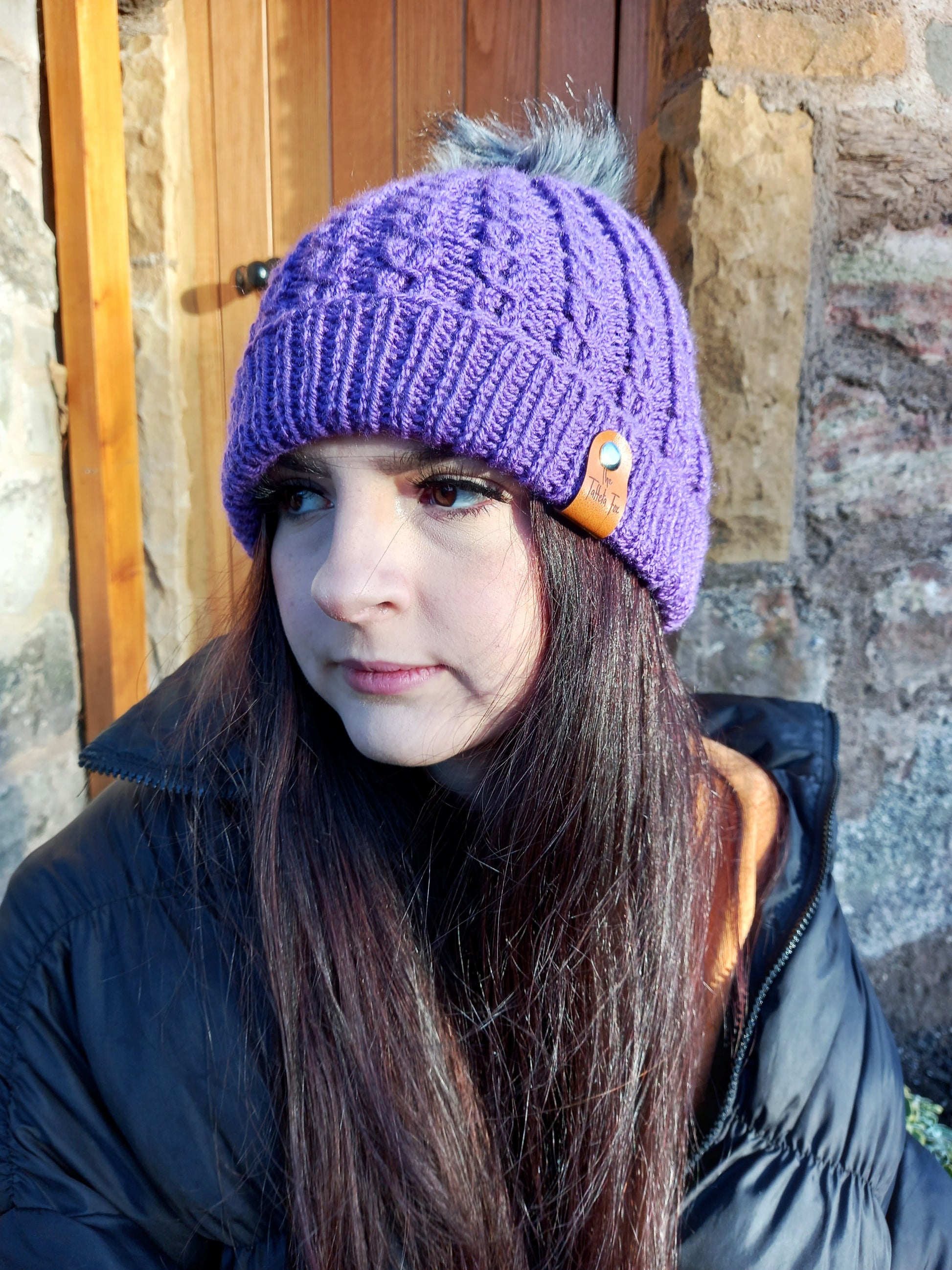 This image shows a young woman with long dark hair wearing a bright purple hand knitted faux cable hat featuring a light grey pompom.