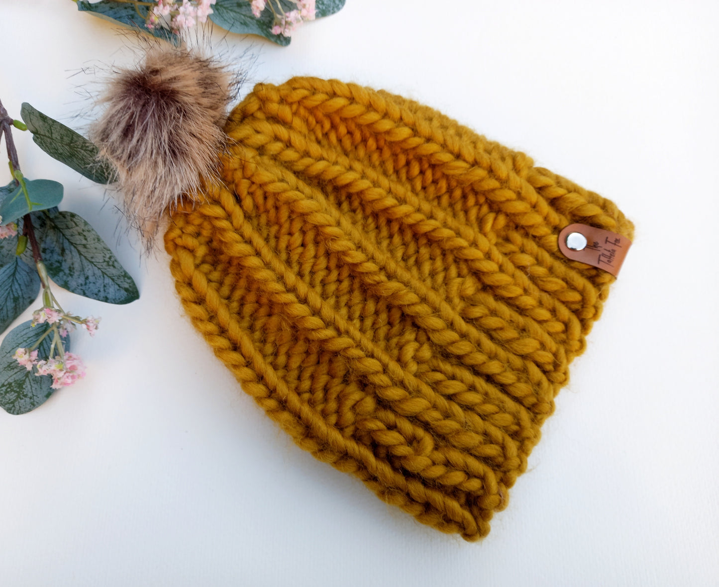 A chunky hand knitted mustard coloured hat lying on a white background with some foliage as decoration. The hat features a brown faux leather tag and brown fluffy faux fur pompom.