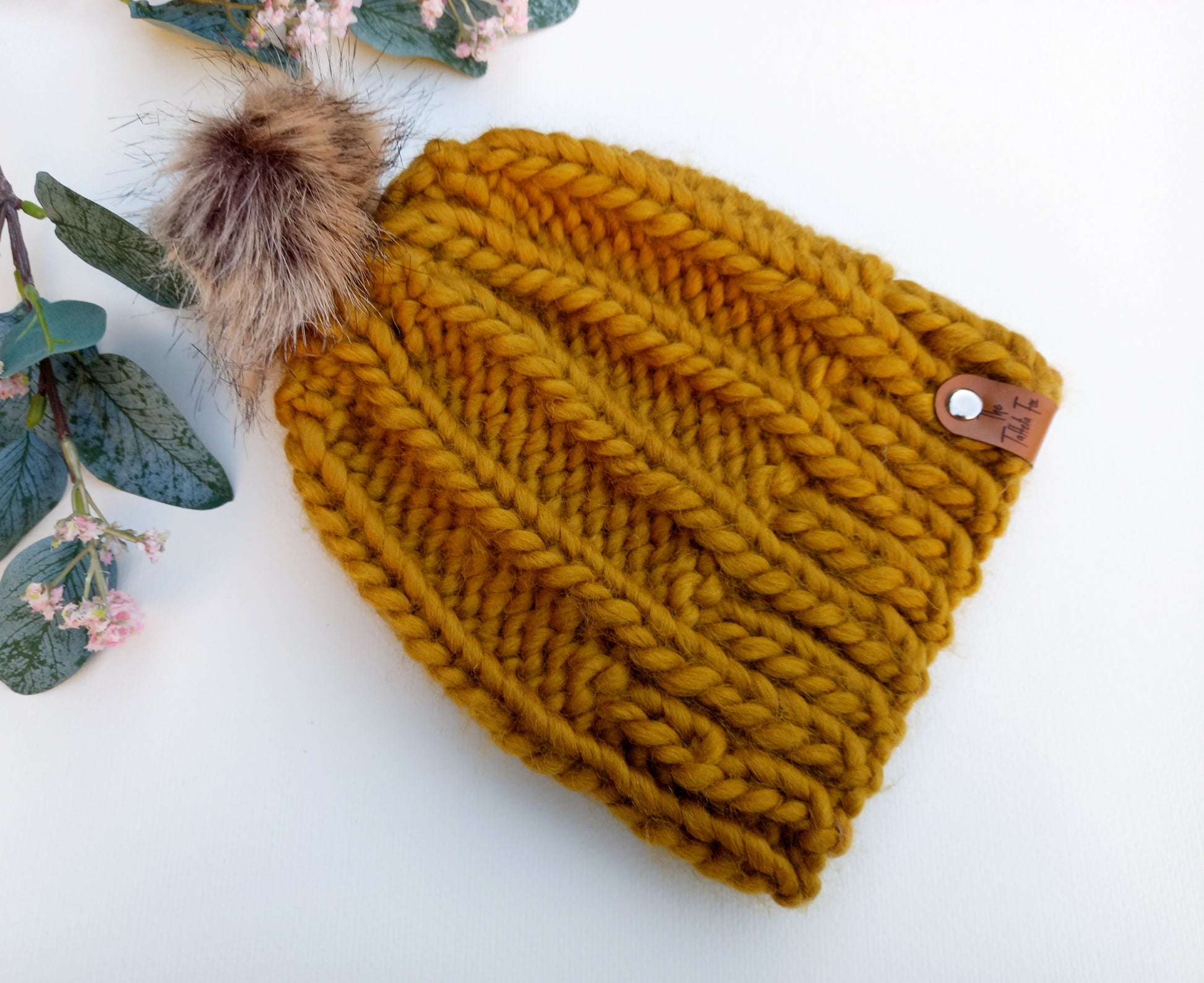 A chunky hand knitted mustard coloured hat lying on a white background with some foliage as decoration. The hat features a brown faux leather tag and brown fluffy faux fur pompom.
