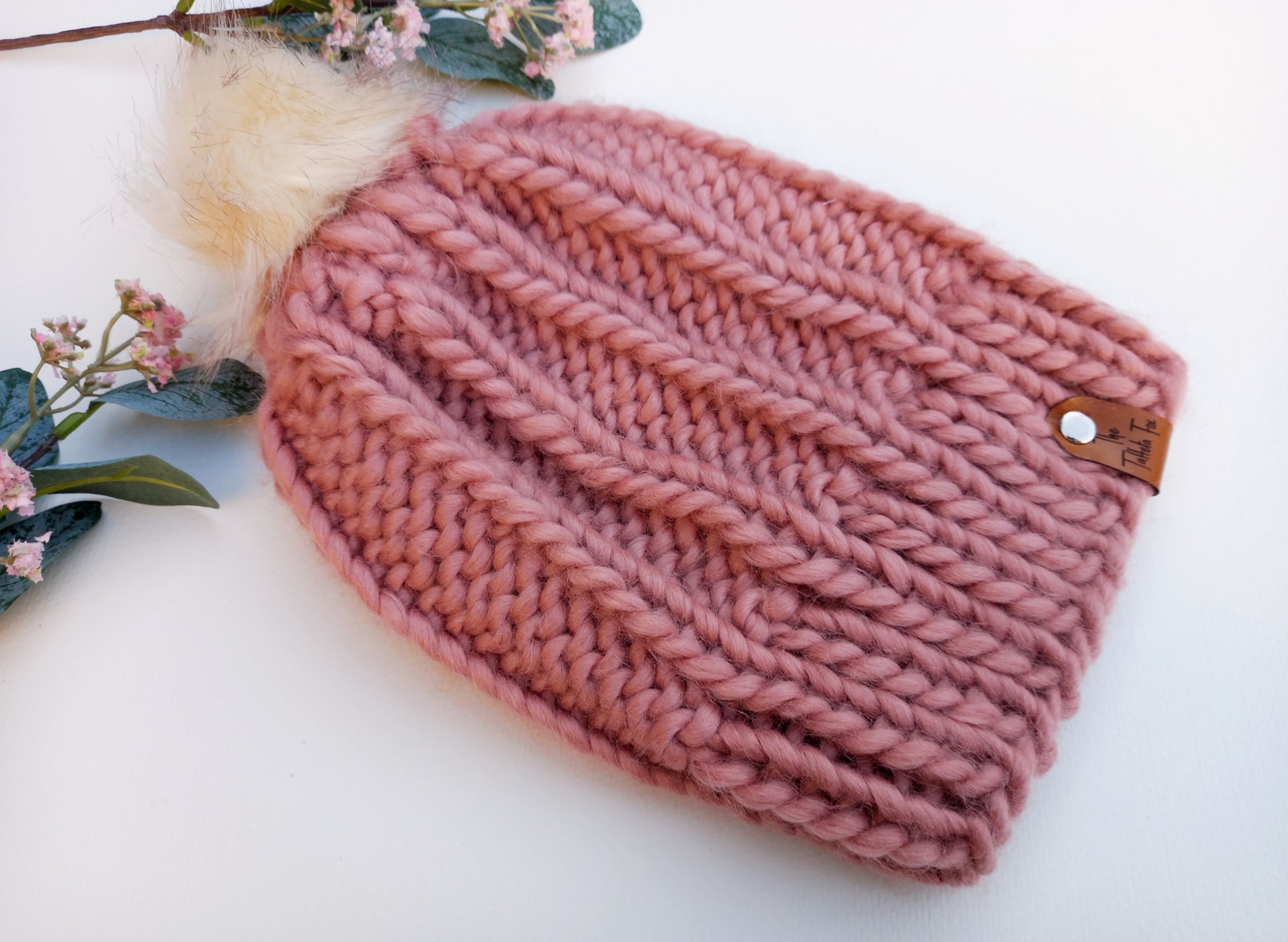 This image shows a hand knitted super chunky hat in pink, lying on a white background with some pink flowers as decoration. The hat features a small brown faux leather tag and creamy coloured pompom.