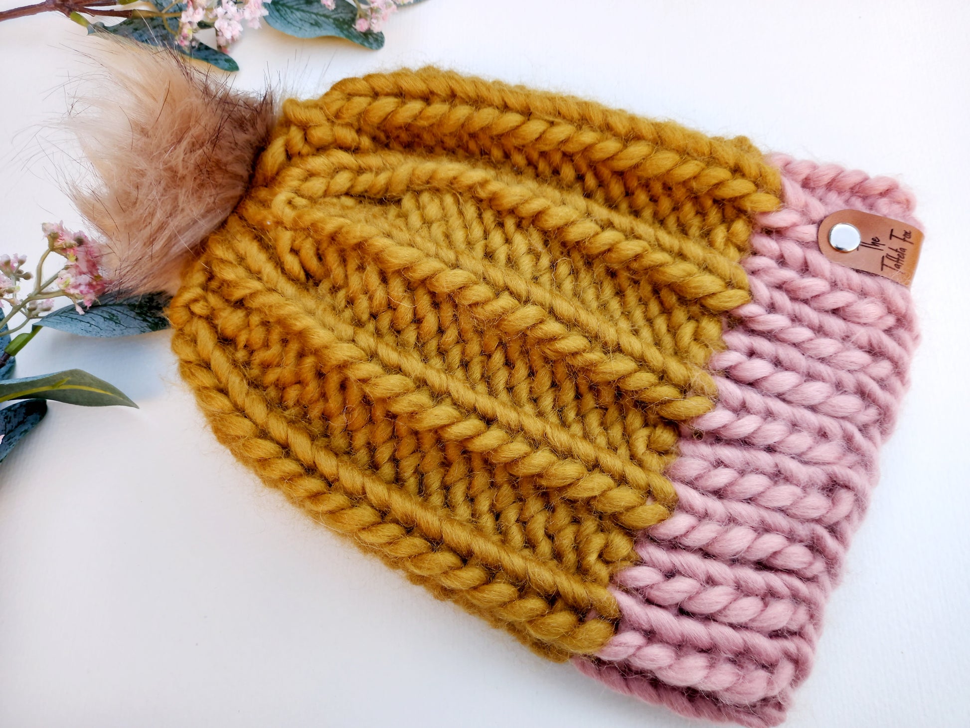 This image shows a super chunky hand knitted hat lying on a white background with some pink flowers as decoration. The hat is mustard with a pink brim and features a beige coloured fluffy pompom and brown faux leather The Taffeta Fox label held on by a small silver screw. 