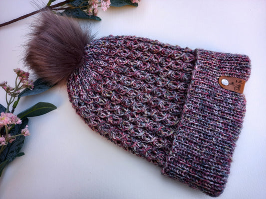 A hand knitted faux cable patterned hat with turned up brim lying on a white background with some foliage as decoration. The hat is in a mix of colours that remind me of Scottish heather and features a brown faux leather tag and fluffy pompom.