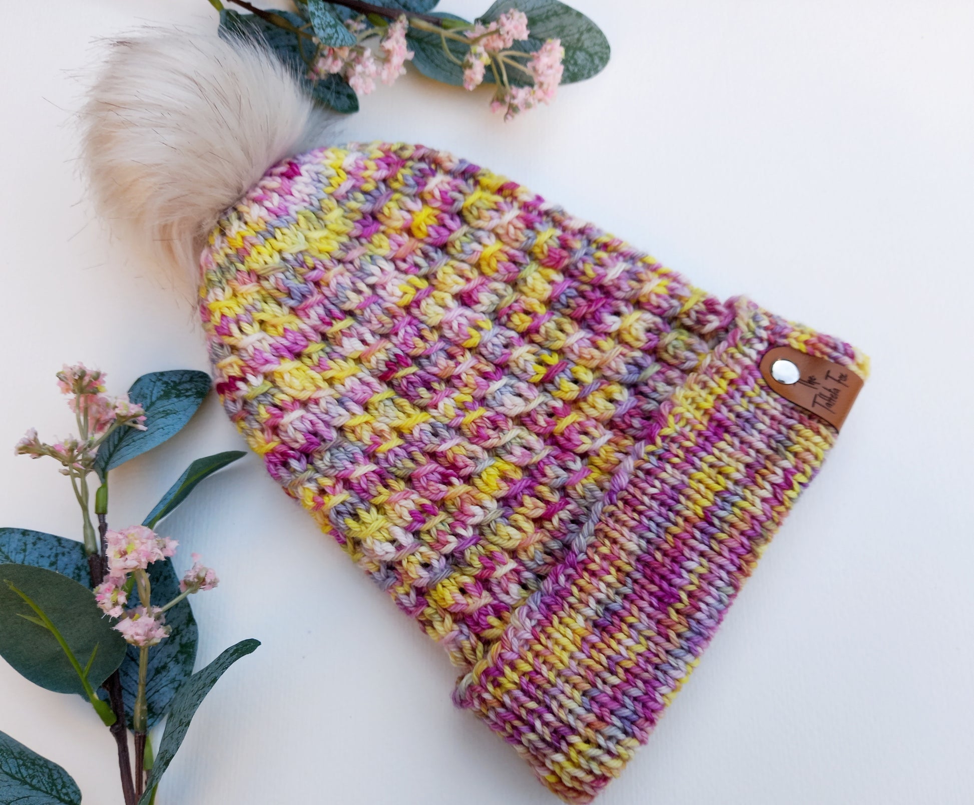 This image shows a hand knitted hat lying on a white background with pink flowers and leaves for decoration. The hat is hand knitted in a faux cable pattern with a wide fold up brim and is a lovely mixture of pinks, yellows, purples and blues. It features a small faux leather 'Taffeta Fox' label and a soft cream removeable pompom.