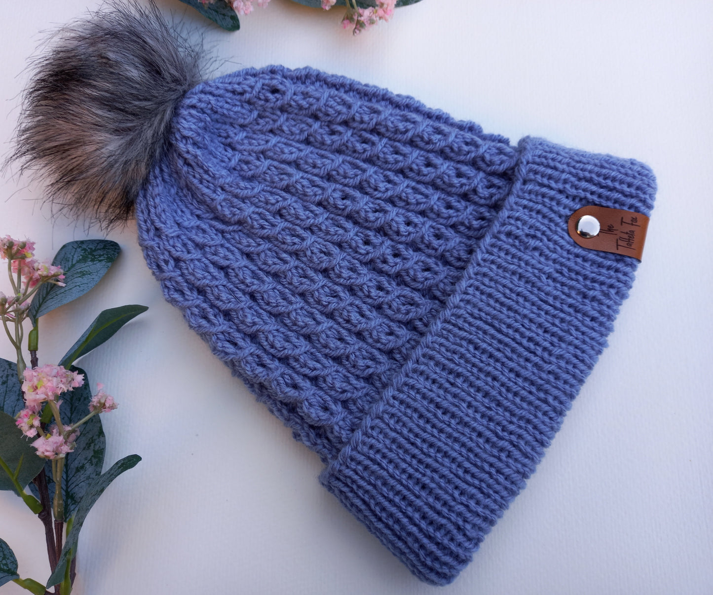 This shows a blue, hand knitted faux cable patterned hat with turned up brim lying on a white background with foliage as decoration. The hat features a brown faux leather The Taffeta Fox tag attached via a silver rivet and a grey pompom.