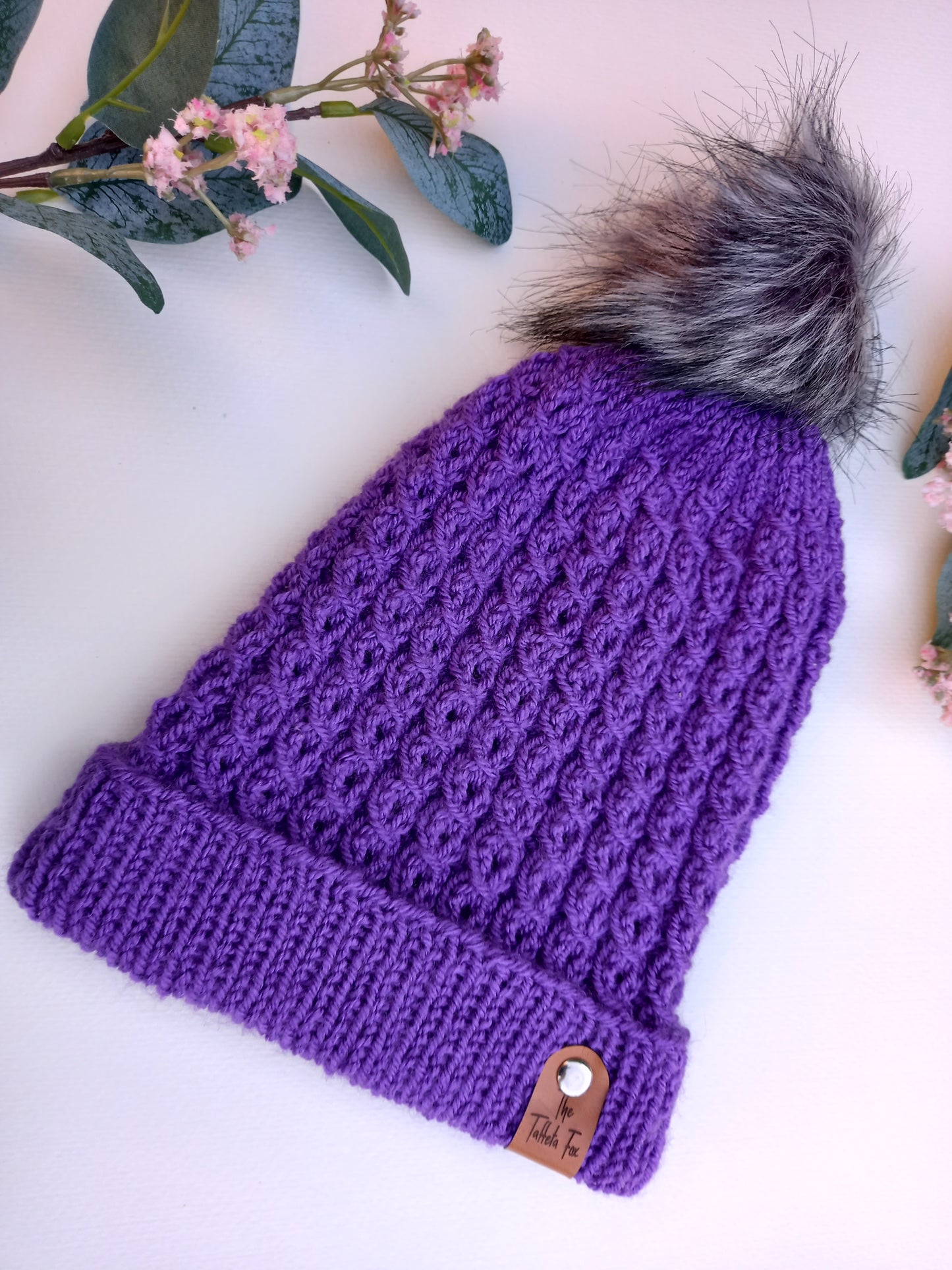 This image shows a bright purple hand knitted faux cable hat lying on a white background some foliage and pink flowers as decoration. The hat features a grey pompom and brown faux leather The Taffeta Fox label attached with a small silver rivet. 